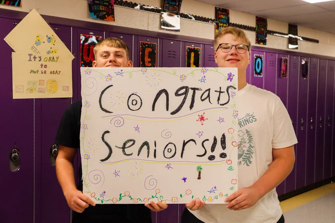 Two students hold a sign reading 'Congrats Seniors'!!' in front of purple lockers
