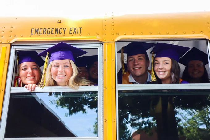 Students in graduation attire sticking their heads out of school bus and smiling