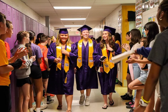 Group of three seniors in graduation attire walk through the Middle School hallway while students hold their hands out for high fives