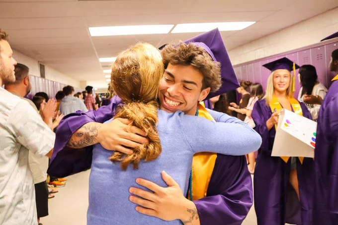 Senior student wearing graduation attire hugs a former teacher in the Middle School hallway