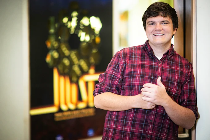 A smiling young man stands leaning against a wall. He wears a red plaid button down and has his hands in front of his chest.