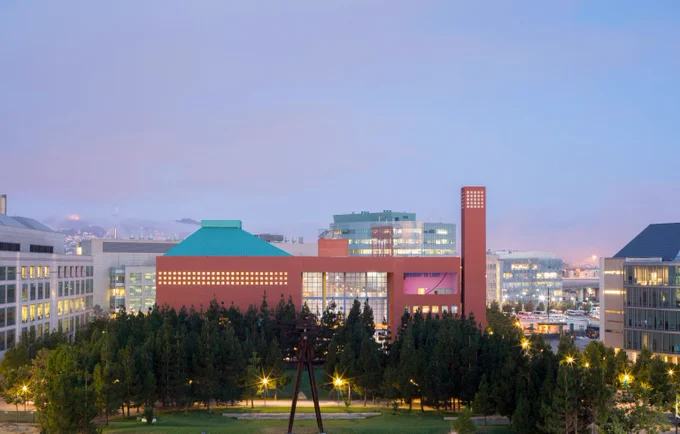 Modern red-brick building with a tall tower, surrounded by trees and city lights at dusk.
