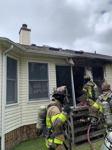Firefighters extinguishing a house fire, with visible damage to the exterior of the building. They are wearing protective gear and using hoses to control the blaze.