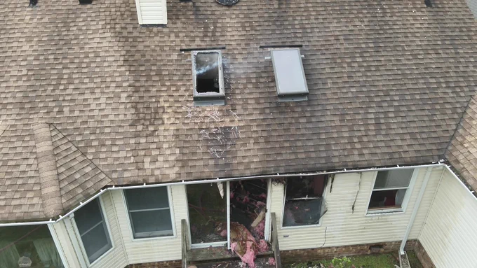 A house with a brown shingle roof shows signs of a fire, including a charred area and a broken window. A skylight is nearby, and smoke residue is visible around the damaged area. The siding below is slightly burned, with pink insulation visible through a broken window.