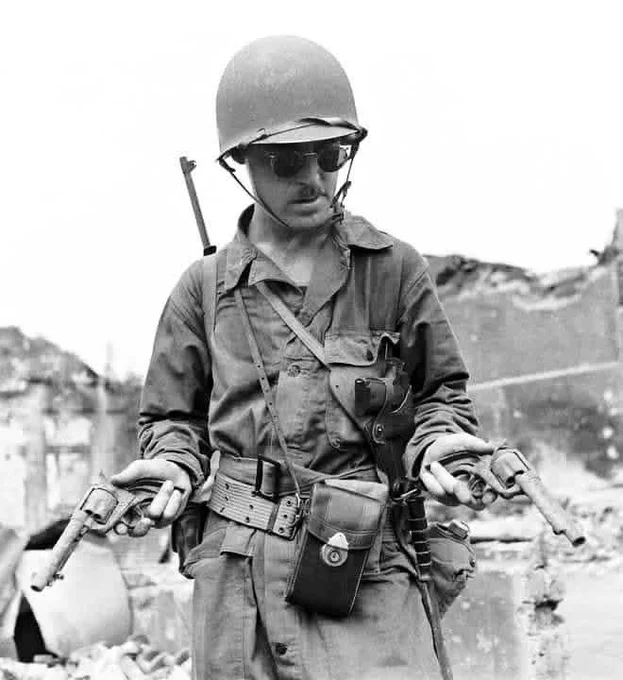 A person in a military uniform and helmet stands holding two pistols amidst a war-torn setting, with damaged buildings in the background.