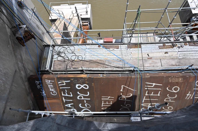 A view looking down into a dewatered spillway tainter gate bay at Robert S. Kerr Lock and Dam. Temporary work platforms and scaffolding surround large steel gate components undergoing repair. Exposed steel plates, cables and maintenance equipment are visible as contractors perform rehabilitation work inside the concrete structure.