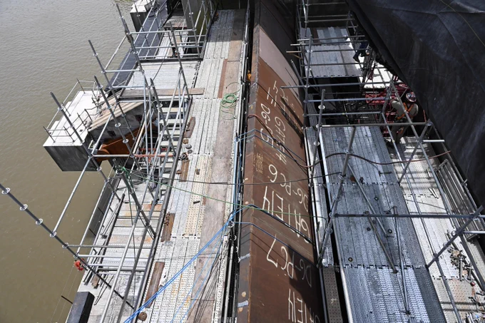 A close-up view of maintenance work on a spillway tainter gate at Robert S. Kerr Lock and Dam. Scaffolding and temporary work platforms surround the large steel gate structure inside a dewatered gate bay. Markings on the gate surface indicate areas identified for repair as part of a rehabilitation project addressing steel deterioration, gate seals, cathodic protection and protective coatings.