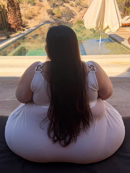 Plus size SSBBW woman seen from behind sitting on a dark sun lounger by an infinity pool in a desert landscape, enormous wide hips and massive round butt completely filling the chair and overflowing on all sides, long dark brown hair cascading down her back, wearing a white swimsuit, sunny desert scenery and pool umbrella visible ahead of her.