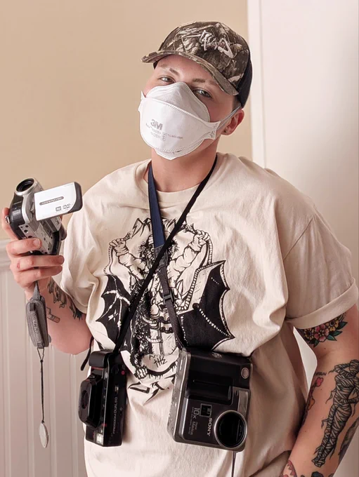 Tank Prusha (@lab_rat_tank), a trans photographer/filmmaker, wearing a 3M Aura N95 respirator, a cap, a T-shirt with graphic, cross-strapped with camera equipment, and holding a handy camcorder with his right hand.