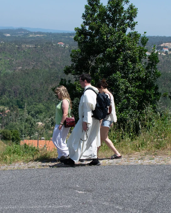 Three people walking along a path with a scenic view of hilly, forested terrain in the background. One person is wearing a long white robe, and another has a backpack. Some houses can be seen among the trees.