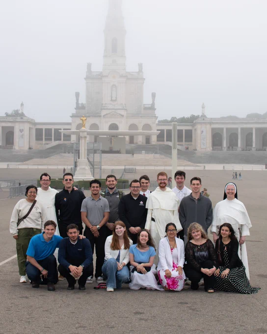 A group of people are posing for a photo in front of the Sanctuary of Fátima. The background shows a foggy view of the basilica. Some members of the group are wearing religious attire.