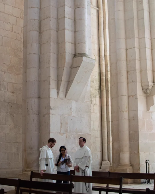 A group of three individuals, including two in long white robes, stand talking inside a grand stone building. The interior features tall pillars and historic architecture, suggesting it might be a church or cathedral.