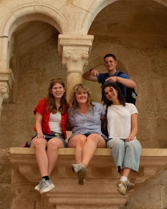 Four people are sitting and standing on a stone ledge with arched architecture in the background. One person is pointing playfully with a smile. They appear to be enjoying a historic setting.