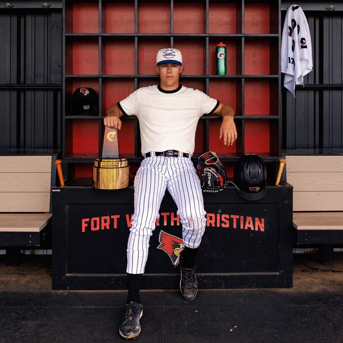 Grady Emerson poses against a Fort Worth Christian helmet and bat rack in a cream t shirt sand white pinstriped pants. He rests his sarm on a Gatorade trophy