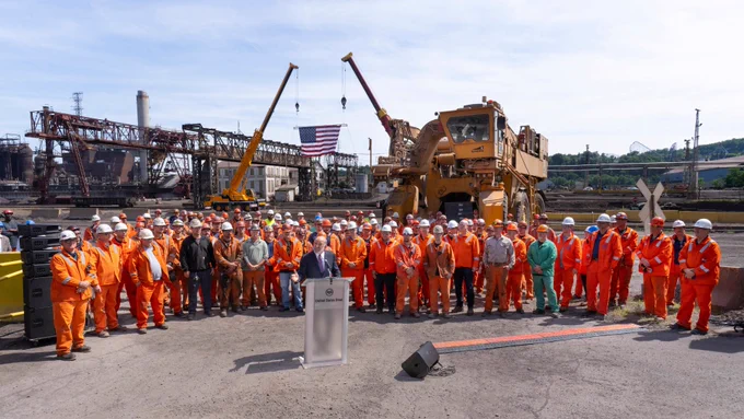 Group of industrial workers in orange safety gear and helmets standing attentively outdoors at Mon Valley Works - Edgar Thomson Plant with heavy machinery and an American flag in the background as Commerce Secretary Lutnick speaks.