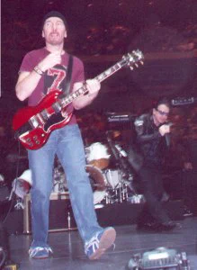 Edge playing Gibson SG guitar while Bono sings into the mic behind him.