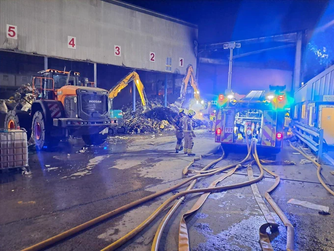 A photo at night showing hoses laid out from a fire engine. Two firefighters are in the middle of the shot, and a large tractor is on the left. The background is a recycling centre.