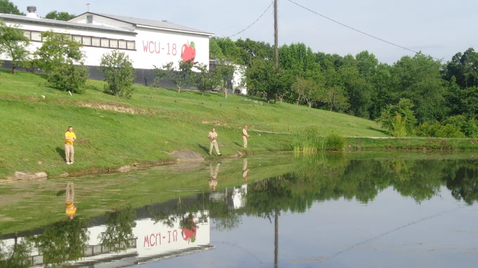 Three people fishing along the grassy bank of a calm pond with a white building and trees in the background.