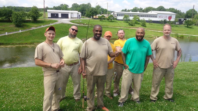 Group of six men standing on grass near a pond, dressed casually in neutral and green tones, with fishing rods and a rural backdrop.