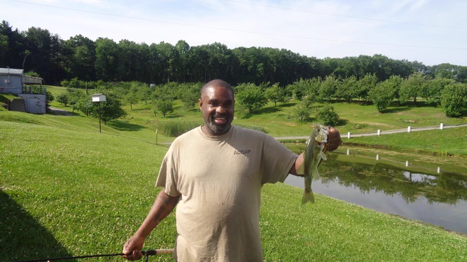Person standing on grassy hill near pond holding a largemouth bass fish by its mouth.