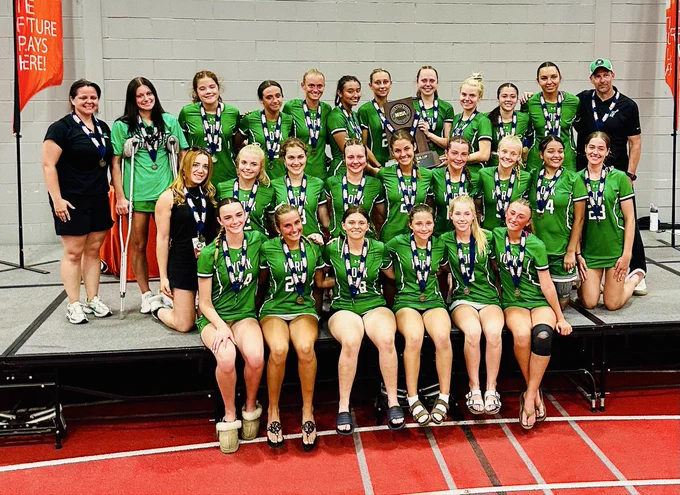 A women's soccer team poses for a group photo on a podium, wearing green jerseys and medals, celebrating their achievement.
