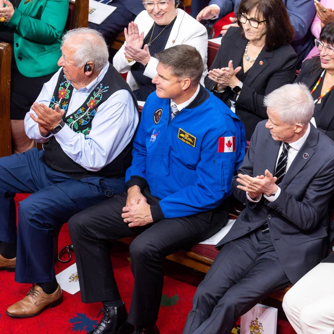 Canadian astronaut Jeremy Hansen sitting in a gallery and clapping, wearing a bright blue flight jacket with a Canadian flag patch.