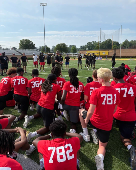 A group of young athletes wearing red numbered shirts gather on a football field, listening attentively to coaches standing under a bright sky.
