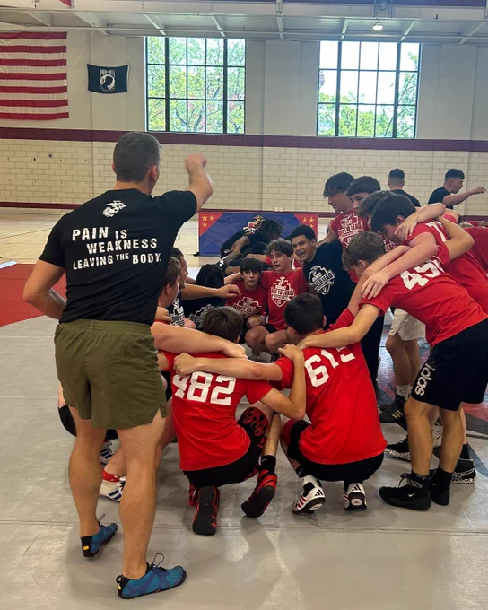 A group of athletes in red jerseys gather for a motivational huddle in a gym. Coaches in black shirts stand nearby. The atmosphere is focused and energetic.