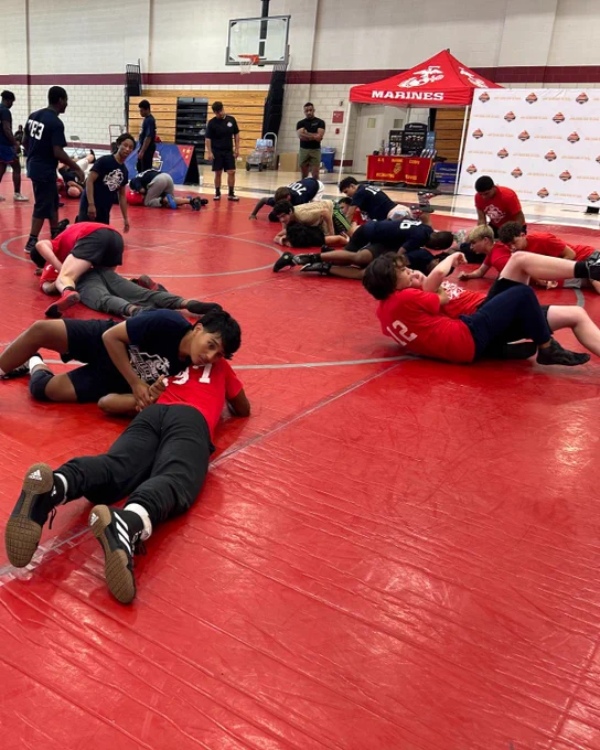 A group of athletes participating in a wrestling practice session on a red mat in a gymnasium. The environment is energetic and focused.