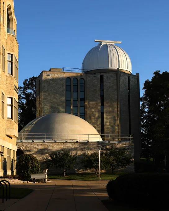 A brick building with large, white domes, part of the Ritter Planetarium and Brooks Observatory, is seen against a clear blue sky. Trees and a walkway surround the observatory.