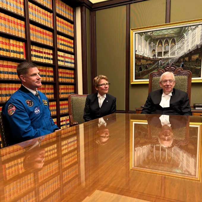 Jeremy Hansen dans son blouson de vol bleu assis à une longue table de conférence en bois aux côtés de deux hauts fonctionnaires en tenue officielle.