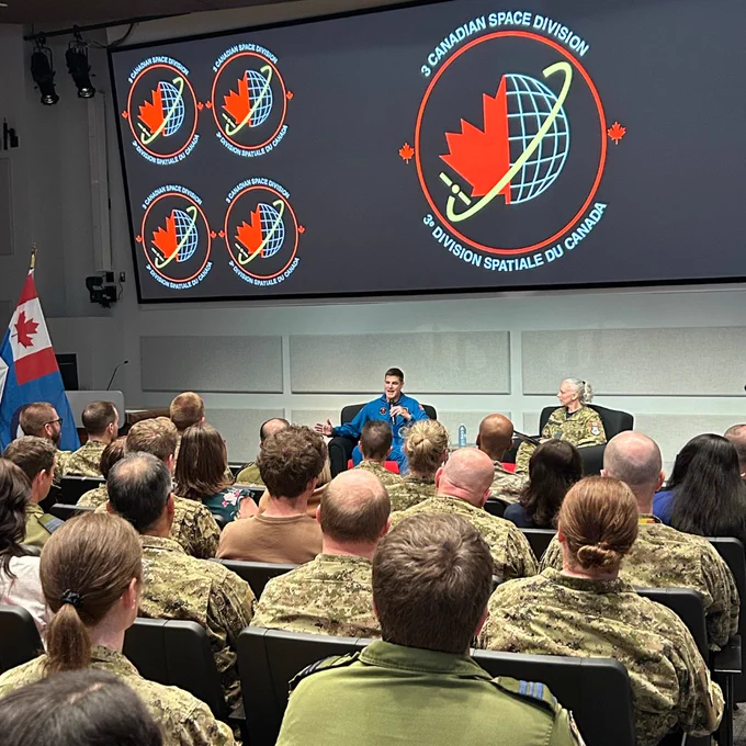 Jeremy Hansen s'exprimant sur scène devant un public de militaires, avec un grand écran affichant le logo de la « 3e Division spatiale du Canada » derrière lui.