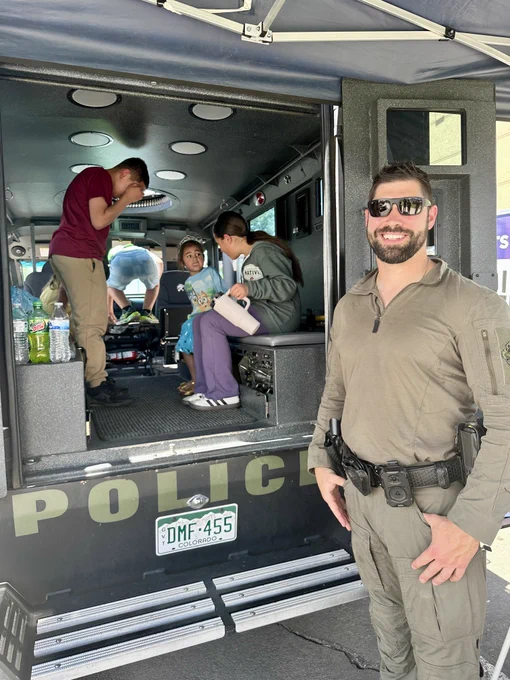 Children exploring the interior of a police vehicle while a uniformed officer stands nearby outside the open door.