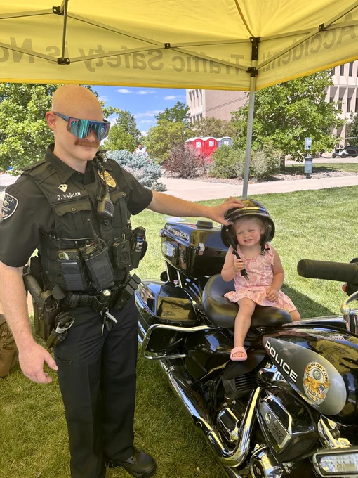 Police officer standing next to a young child sitting on a black police motorcycle under a yellow tent on a sunny day.