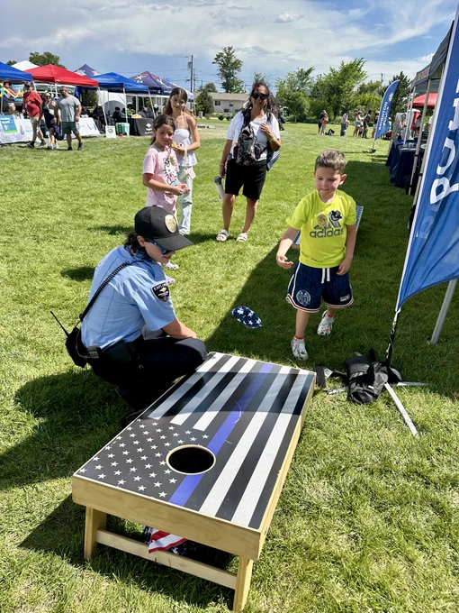 Child playing cornhole with a police officer at an outdoor community event on a sunny day.