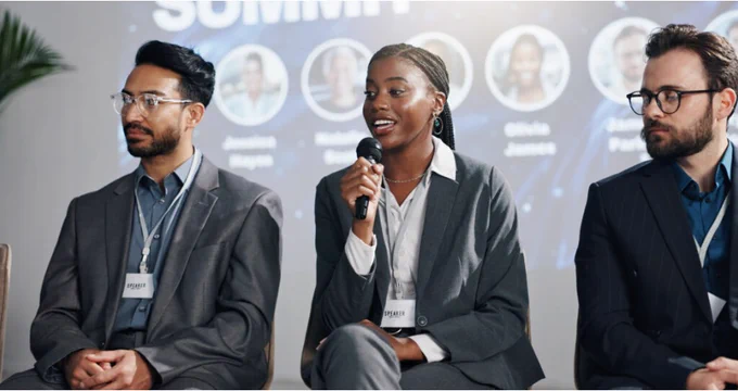 Three business professionals seated on a panel during a conference discussion. The woman in the center speaks into a handheld microphone while the two men on either side listen attentively. All three wear business attire and conference badges, with a blurred event backdrop and speaker headshots behind them.