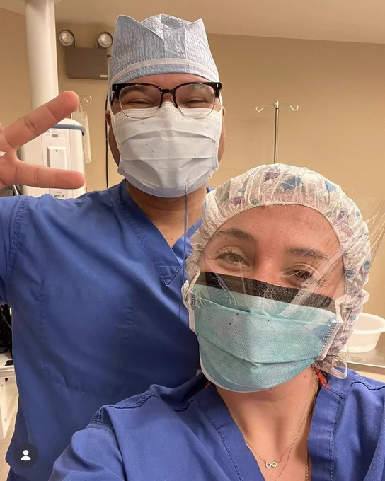 Two healthcare professionals in blue scrubs and surgical masks pose in a hospital setting. One person makes a peace sign while the other smiles visibly under a transparent face shield.
