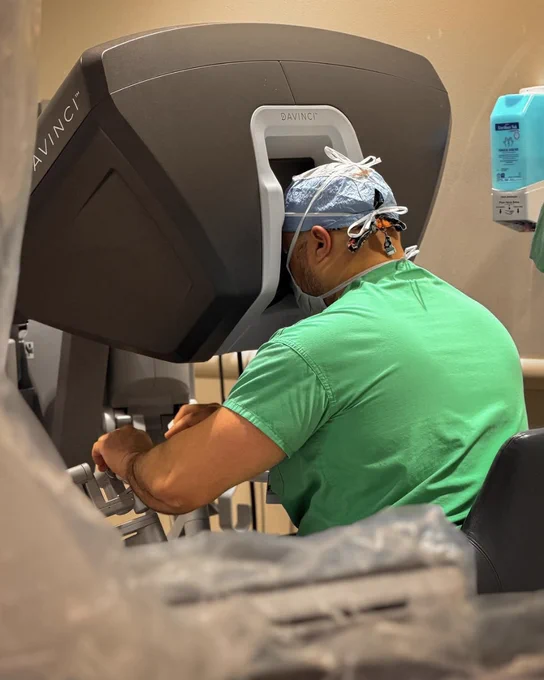 A person in medical scrubs is using a Da Vinci surgical robot console, focused on the controls.