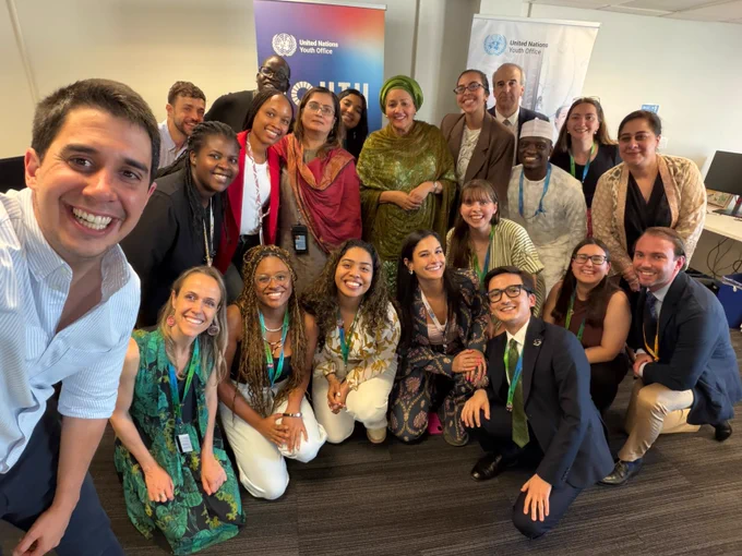 A group of diverse staff at the United Nations Youth Office posing together for a selfie with the UN Deputy Secretary-General at the United Nations Youth Office event indoors.