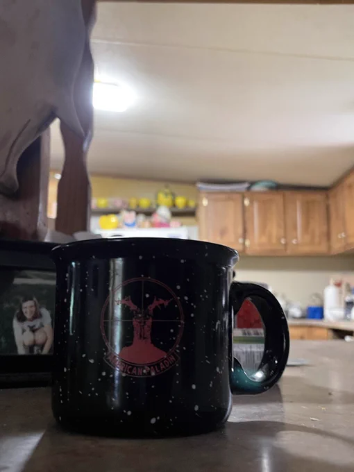 Black ceramic mug on a countertop.  
The mug is cylindrical with slightly flared sides and a rounded rim. It is mostly black with white speckled dots scattered over the surface, giving it an enamel-style look. On the front there is a red circular logo: inside the circle is an outline of the Upper Peninsula of Michigan with crosshairs over it, like a scope reticle. Around the lower curve of the circle, partially readable, are red letters that appear to say “AMERICAN PALADIN” or something very similar. The handle is large and solid, also black with some light reflecting along its edges.