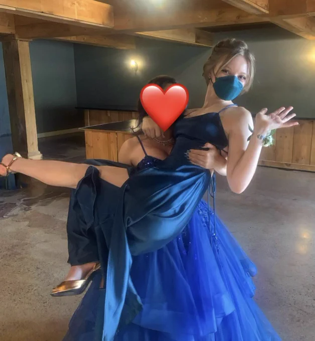 A young woman with short blonde hair, a blue respirator, and a matching blue satin slip dress is being carried by another young woman in a royal blue full-skirted tulle dress.