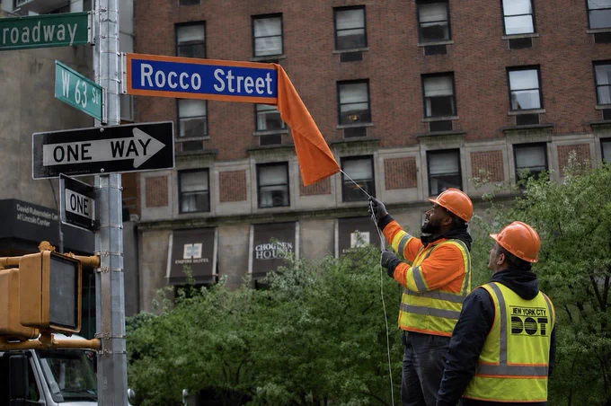 An altered photo showing two N Y C D O T workers unveiling an orange and blue Rocco Street sign. Serves him right.