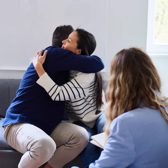 A couple embracing each other in a hug, with a therapist in the foreground. 