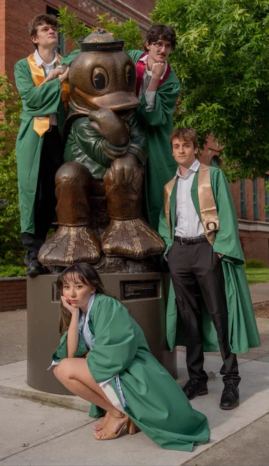 A group of people in green graduation robes pose around a statue of the University of Oregon's mascot, The Duck, outside a building.