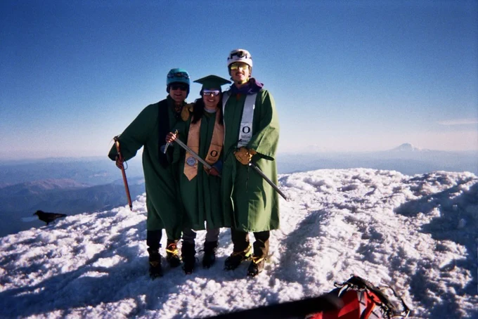 Three individuals, wearing green graduation gowns and caps, stand together on a snow-covered mountain summit. They are holding ice axes, suggesting a climbing expedition. The sky is clear, and distant mountains are visible in the background.