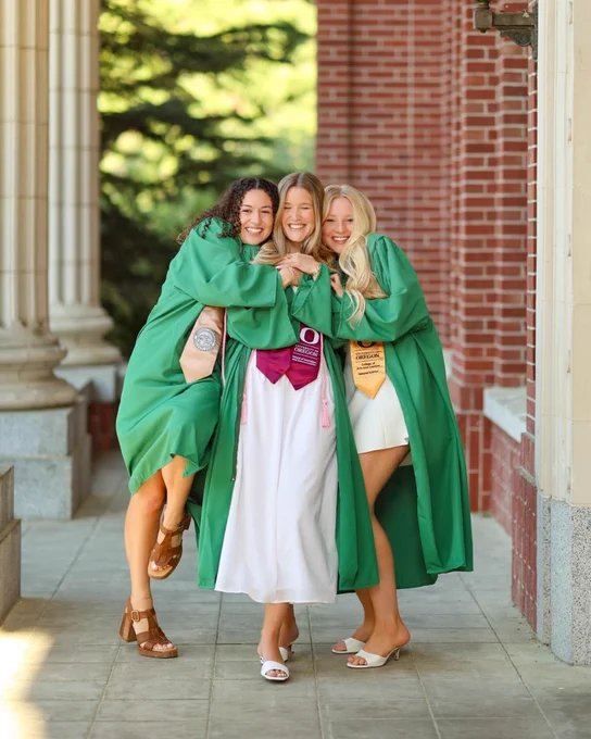 Three people wearing green graduation gowns stand close together, smiling and embracing on a stone walkway. They have various colorful stoles and cords. A brick wall and columns are in the background.