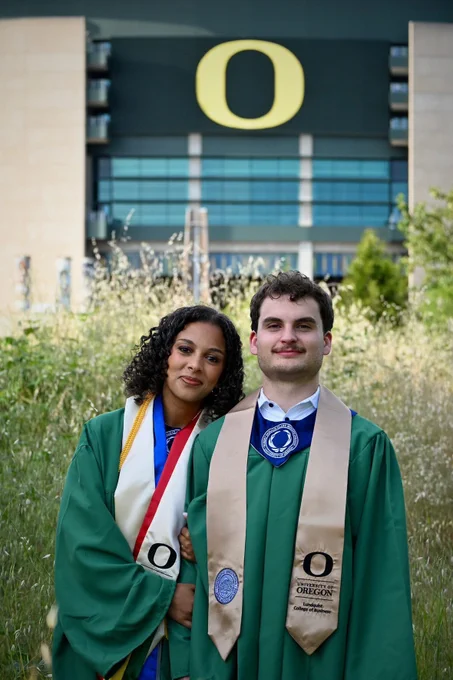 Two individuals in graduation attire stand together in front of Autzen Stadium with the University of Oregon logo. Both wear green robes and sashes. Grass and greenery surround them.