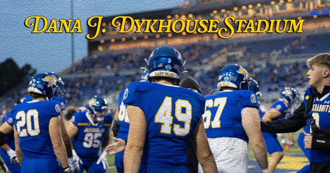 Football players in blue uniforms with yellow accents gather near the sidelines. Stylized text stating "Dana J. Dykhouse Stadium" is prominently displayed at the top. A crowd is visible in the stadium seats.