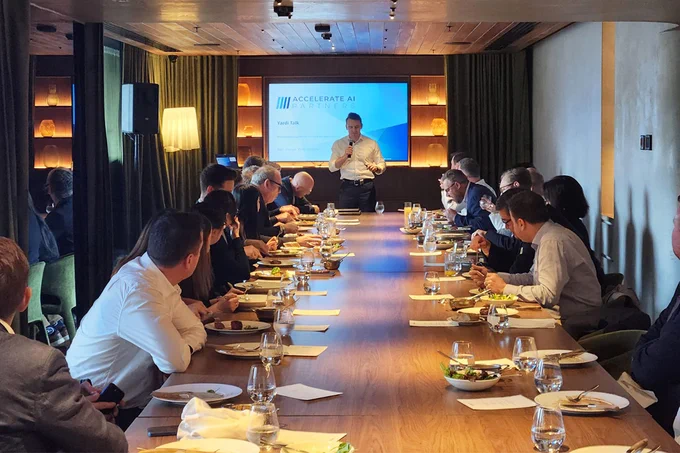 A group of people seated around a long table in a conference room, listening to a presentation. A person stands at the front near a screen displaying "Accelerate AI" and "Fireside Chat." Plates of food and drinks are on the table.