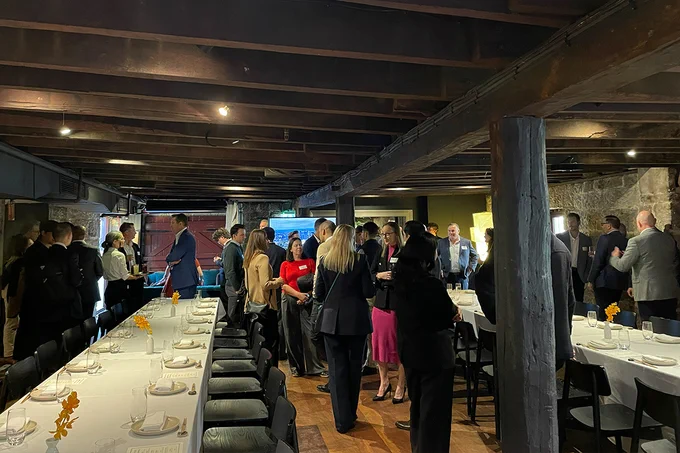 A group of people are gathered in a rustic dining room with wooden beams and stone walls. They are standing and conversing near tables set for a meal, with white tablecloths and neatly arranged place settings.